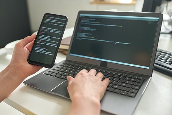 manos de un programador sosteniendo un celular y tecleando en la computadora sobre su escritorio