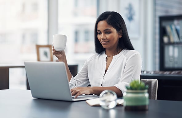 mujer que trabaja en su oficina con una laptopn mientras sonrie al sostener una taza de cafe
